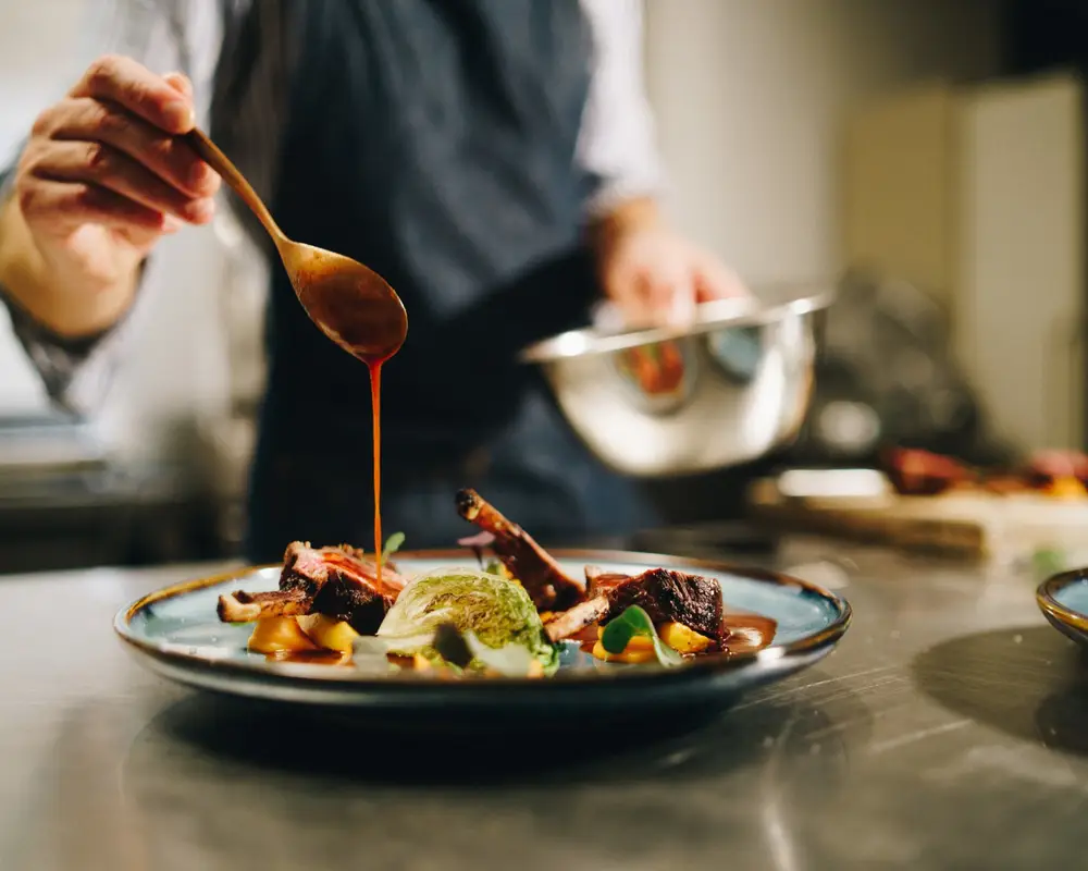Chef emplatando: vierte salsa con una cuchara sobre un plato de carne y verduras
